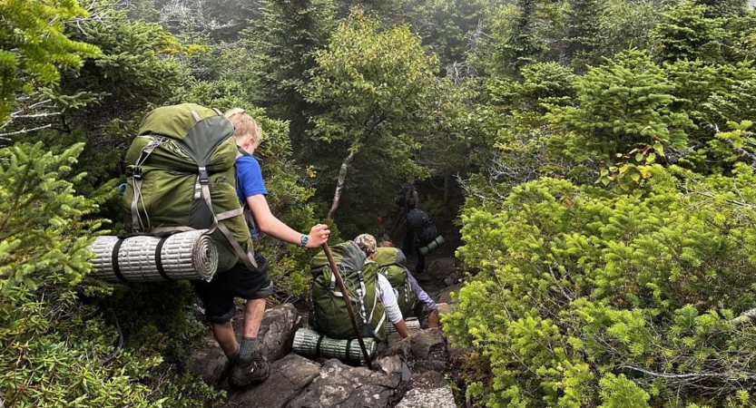 A group of students carrying backpacks maneuver down an incline in a green wooded area. 